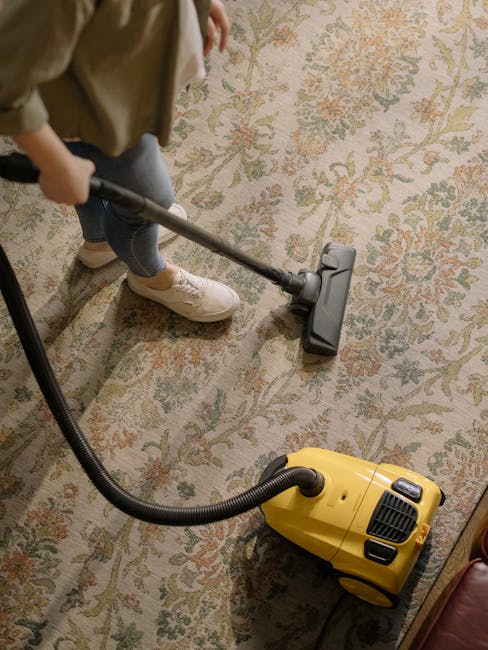 A person using a yellow and black vacuum cleaner with a long hose attachment to deep clean a patterned carpet in a residential setting. The individual's legs are visible, wearing light-colored sneaker