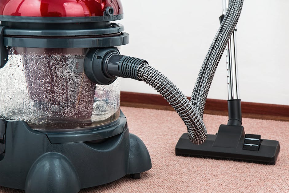 A professional cleaner dressed in a white protective suit, face mask, and orange gloves is operating a vacuum cleaner in a modern, well-lit domestic setting. The room features smooth light grey tiled