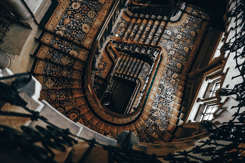 An aerial view of a spiral staircase inside a period property, featuring wooden banisters with intricate black wrought iron balustrades. The staircase is covered by a patterned carpet runner with flor